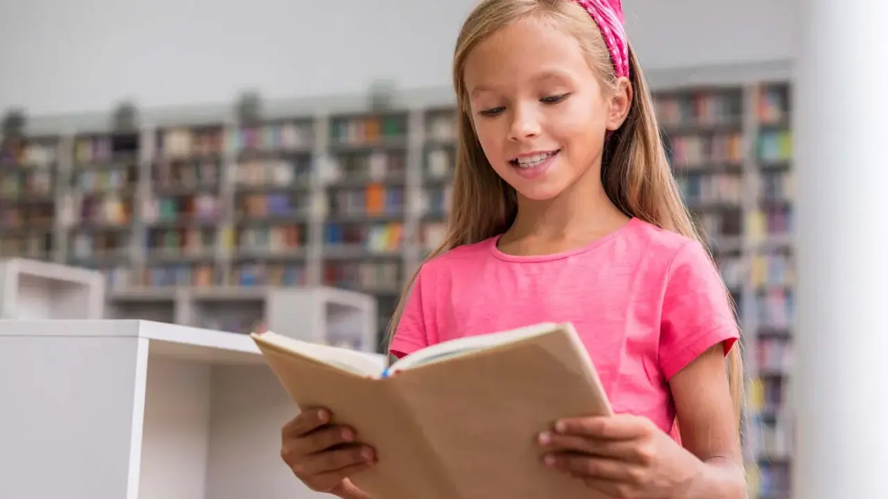 Ein Mädchen mit pinkem Haarband liest in der Schulbibliothek. Ein Beispiel für Leseförderung in der Grundschule, das Freude am Buch zeigt.