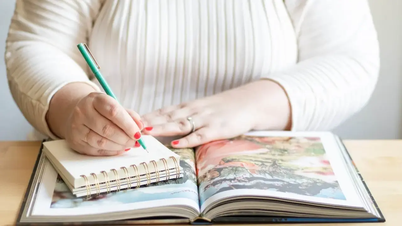 Frau schreibt mit grünem Stift in Notizbuch, während sie ein aufgeschlagenes Fotoalbum betrachtet. Sie erstellt ihr persönliches Jahrbuch.