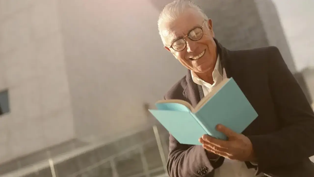 Ein älterer Mann mit Brille lächelt und hält ein hellblaues Buch. Er scheint Freude am Auswendiglernen von Gedichten zu haben.
