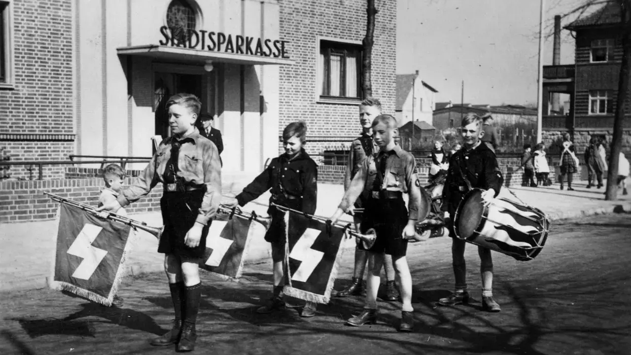 Jungen in Uniformen marschieren mit Fahnen und Trommel vor der Stadtsparkasse. Ein Bild der vermeintlichen Volksgemeinschaft.