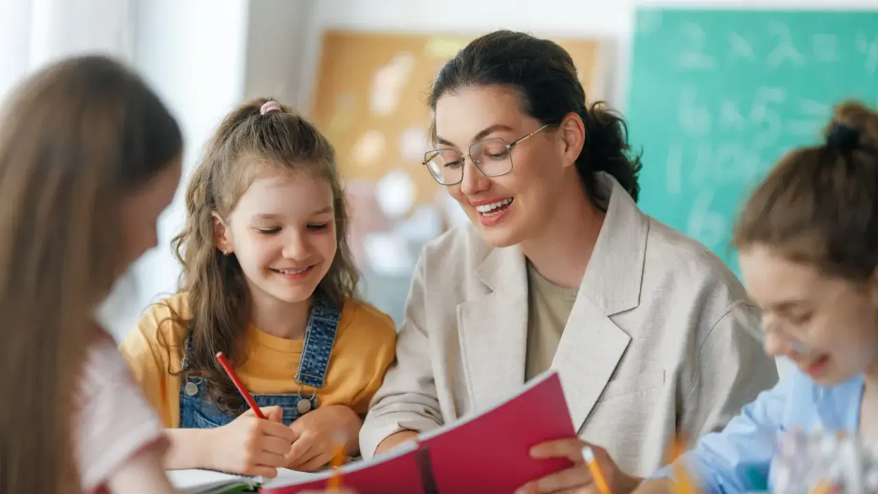 Lehrerin erklärt Kindern etwas aus einem Buch. Eine Tafel mit Rechenaufgaben im Hintergrund deutet auf die Bedeutung von Bildungspolitik für die Zukunft hin.