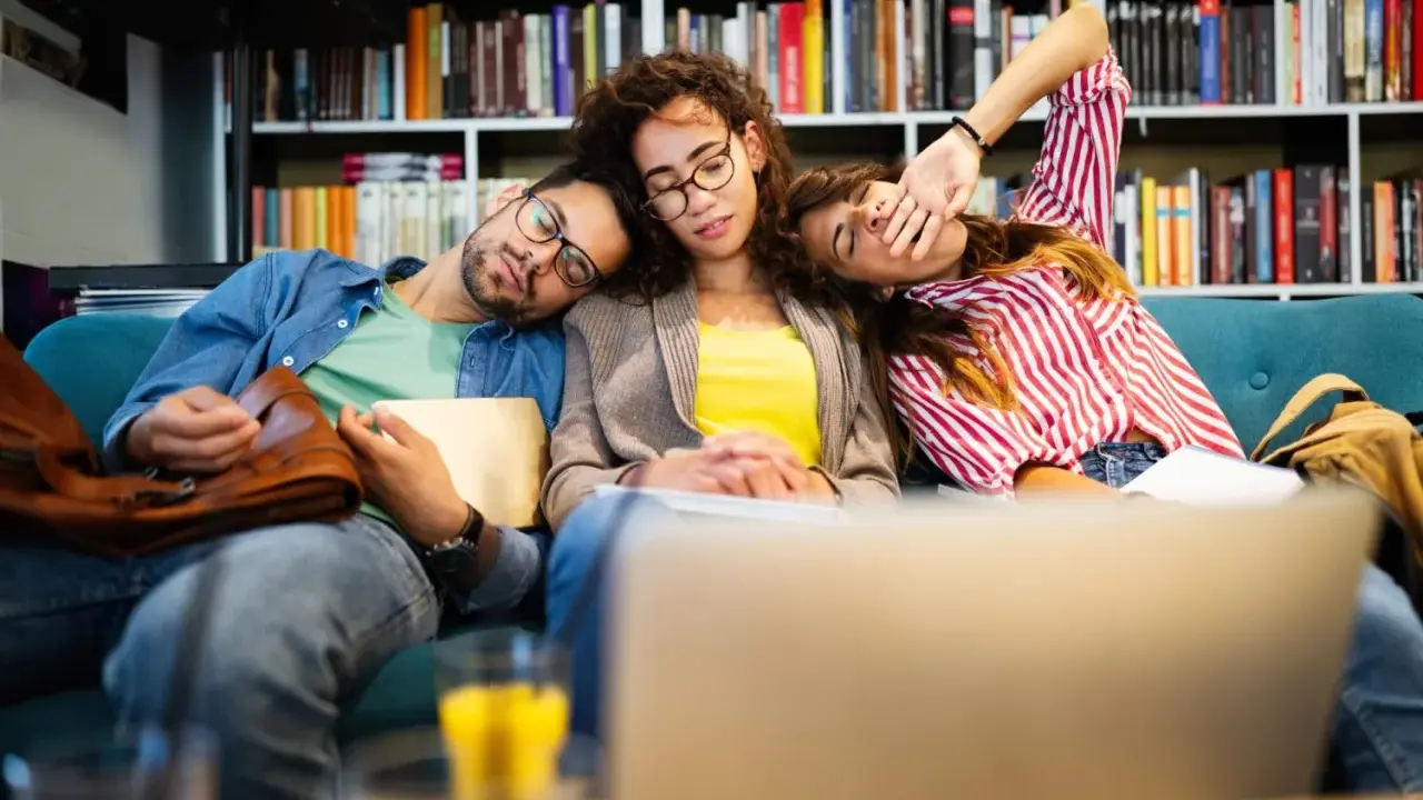 Drei erschöpfte Studenten schlafen auf einer Couch, umgeben von Büchern. Der abi deutsch-Test war wohl zu anstrengend.