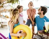 Familie lacht am Strand, genießt die Sommerferien. Ein gelber Schwimmring und Cocktails deuten auf Urlaubsfreude in den Bundesländern hin.