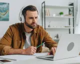 Mann mit Kopfhörern lernt online, nutzt Laptop und Notizbuch. Er wirkt konzentriert, während er mit einem Stift tippt. Ein typisches Bild für das heutige "Home-Office"-Zeitalter, das viele Anglizismen mit sich bringt.
