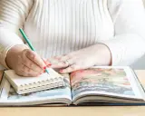 Frau schreibt mit grünem Stift in Notizbuch, während sie ein aufgeschlagenes Fotoalbum betrachtet. Sie erstellt ihr persönliches Jahrbuch.