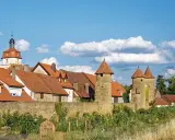 Eine malerische **Stadt im Mittelalter** mit roten Dächern, steinernen Türmen und einer alten Stadtmauer unter blauem Himmel.