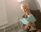 Ein älterer Mann mit Brille lächelt und hält ein hellblaues Buch. Er scheint Freude am Auswendiglernen von Gedichten zu haben.