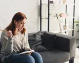 Junge Frau mit roten Haaren sitzt auf einem Sofa und schreibt in ein Notizbuch. Sie hält einen Bleistift, als würde sie über das richtige Metrum für ein Gedicht nachdenken.