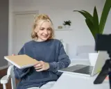 Junge Frau gibt online Nachhilfe, hält ein Notizbuch und lächelt in die Kamera.