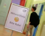Schild mit der Bezeichnung "Abiturprüfung. Bitte Ruhe!" und einem Emoji, das zum Schweigen auffordert. Ein Schüler geht im Hintergrund vorbei.