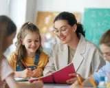Lehrerin erklärt Kindern etwas aus einem Buch. Eine Tafel mit Rechenaufgaben im Hintergrund deutet auf die Bedeutung von Bildungspolitik für die Zukunft hin.