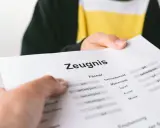 Ein Zeugnis wird überreicht. Die Noten sind gemischt, von "gut" bis "befriedigend", aber keine "ungenügend" Note.