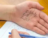 Formeln auf der Handfläche, um in der Schule zu spicken. Ein Schüler schreibt physikalische Gleichungen auf seine Hand, während er in sein Heft schaut.
