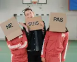 Drei Personen mit Pappkartons auf dem Kopf, auf denen "NE", "SUIS" und "PAS" steht. Sie bilden die französische Verneinung "ne suis pas".