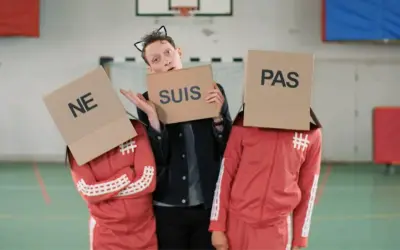 Drei Personen mit Pappkartons auf dem Kopf, auf denen "NE", "SUIS" und "PAS" steht. Sie bilden die französische Verneinung "ne suis pas".