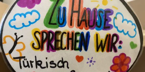 Bunte Zeichnung mit der Aufschrift "Zu Hause sprechen wir: Türkisch & Deutsch". Ein Beispiel für vorurteilsbewusste Pädagogik, die Mehrsprachigkeit feiert.