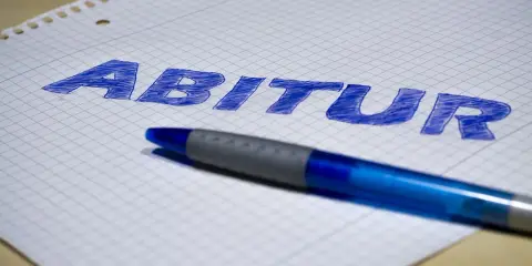 Blauer Stift liegt auf kariertem Papier, auf dem das Wort "Abitur" geschrieben ist. Der **Abitur**-Abschluss rückt näher.