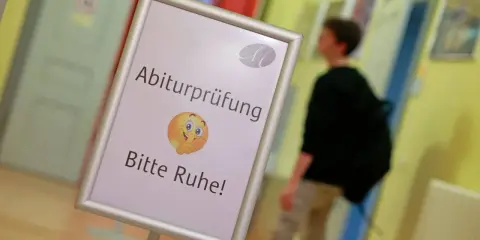 Schild mit der Bezeichnung "Abiturprüfung. Bitte Ruhe!" und einem Emoji, das zum Schweigen auffordert. Ein Schüler geht im Hintergrund vorbei.