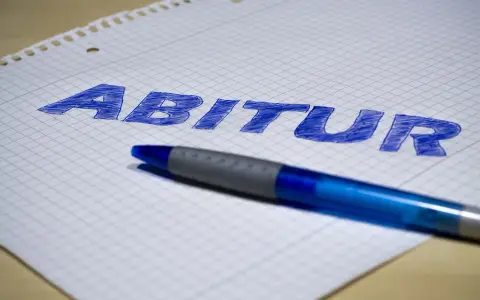 Blauer Stift liegt auf kariertem Papier, auf dem das Wort "Abitur" geschrieben ist. Der **Abitur**-Abschluss rückt näher.