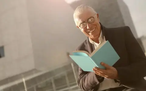 Ein älterer Mann mit Brille lächelt und hält ein hellblaues Buch. Er scheint Freude am Auswendiglernen von Gedichten zu haben.
