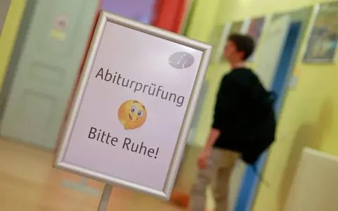 Schild mit der Bezeichnung "Abiturprüfung. Bitte Ruhe!" und einem Emoji, das zum Schweigen auffordert. Ein Schüler geht im Hintergrund vorbei.