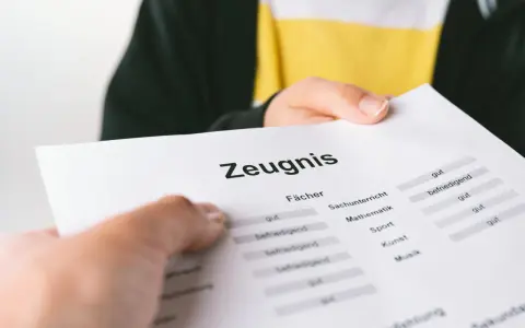 Ein Zeugnis wird überreicht. Die Noten sind gemischt, von "gut" bis "befriedigend", aber keine "ungenügend" Note.