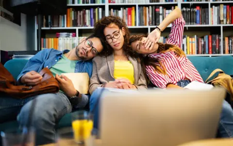 Drei erschöpfte Studenten schlafen auf einer Couch, umgeben von Büchern. Der abi deutsch-Test war wohl zu anstrengend.