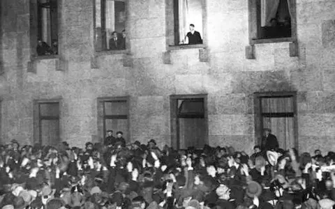 Menge jubelt vor Gebäude, während ein Mann auf einer Leiter steht. Die Szene erinnert an die Zeit des Ermächtigungsgesetzes.