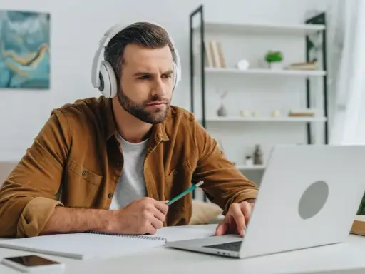 Mann mit Kopfhörern lernt online, nutzt Laptop und Notizbuch. Er wirkt konzentriert, während er mit einem Stift tippt. Ein typisches Bild für das heutige "Home-Office"-Zeitalter, das viele Anglizismen mit sich bringt.