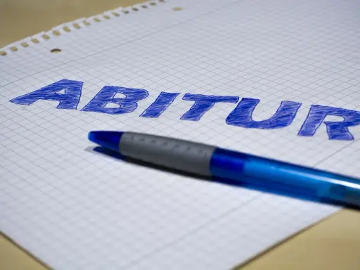 Blauer Stift liegt auf kariertem Papier, auf dem das Wort "Abitur" geschrieben ist. Der **Abitur**-Abschluss rückt näher.