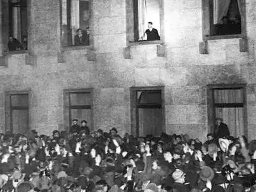 Menge jubelt vor Gebäude, während ein Mann auf einer Leiter steht. Die Szene erinnert an die Zeit des Ermächtigungsgesetzes.