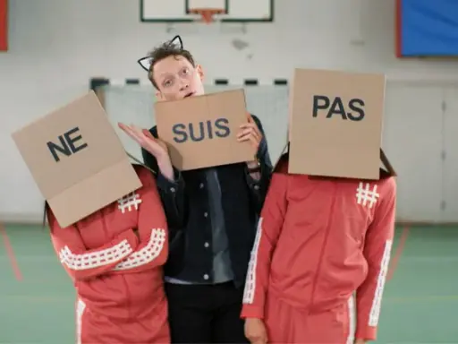 Drei Personen mit Pappkartons auf dem Kopf, auf denen "NE", "SUIS" und "PAS" steht. Sie bilden die französische Verneinung "ne suis pas".