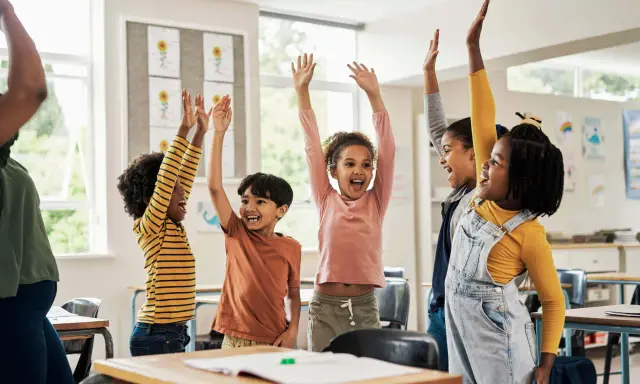 Fröhliche Kinder mit erhobenen Armen in einem Klassenzimmer. Sie nehmen begeistert an Kennenlernspielen für die Grundschule teil.