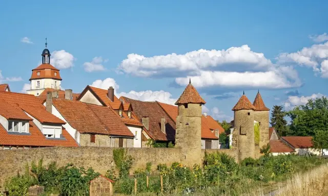 Eine malerische **Stadt im Mittelalter** mit roten Dächern, steinernen Türmen und einer alten Stadtmauer unter blauem Himmel.