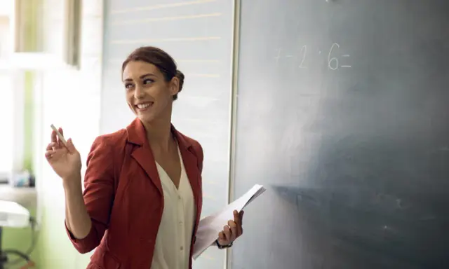 Lehrerin mit Kreide und Unterlagen vor einer Tafel mit einer Matheaufgabe. Sie lächelt und wirkt engagiert, was auf einen interaktiven Unterricht hindeutet.