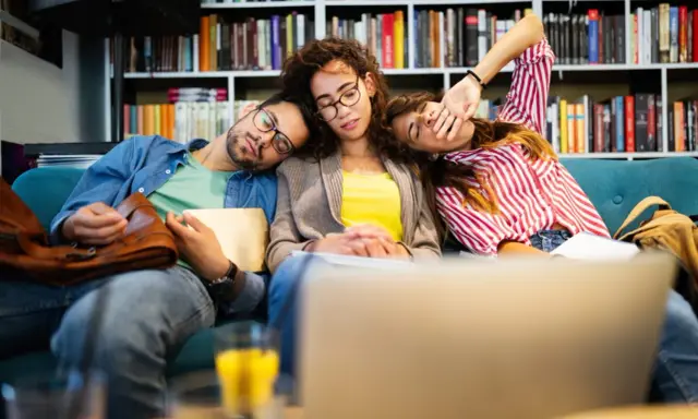 Drei erschöpfte Studenten schlafen auf einer Couch, umgeben von Büchern. Der abi deutsch-Test war wohl zu anstrengend.