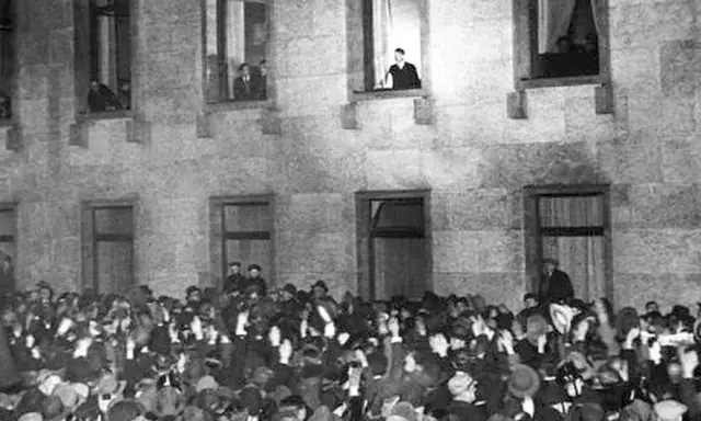 Menge jubelt vor Gebäude, während ein Mann auf einer Leiter steht. Die Szene erinnert an die Zeit des Ermächtigungsgesetzes.