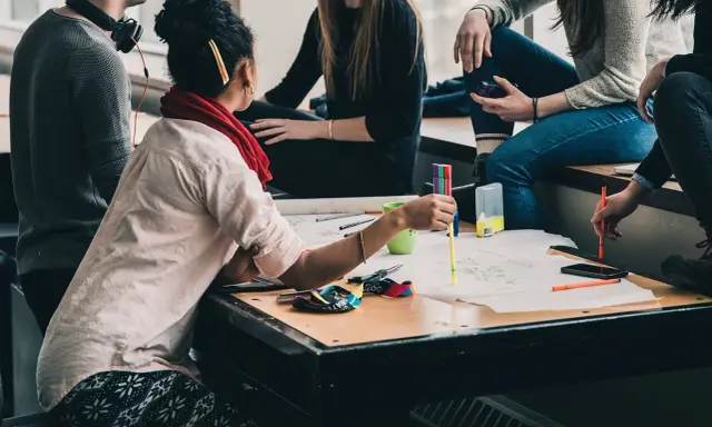 Eine Gruppe junger Leute arbeitet an einem Projekt. Eine Frau mit **Lernschwäche** hält einen Stift und erklärt etwas.