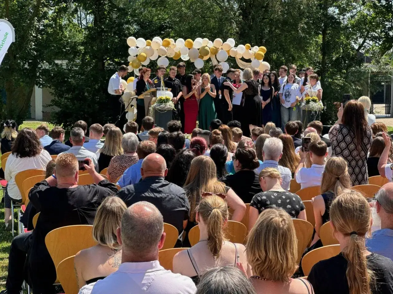 Abschlussfeier der 10. Klasse mit Ballons und feierlicher Stimmung. Viele junge Menschen und Gäste verfolgen die Zeremonie.