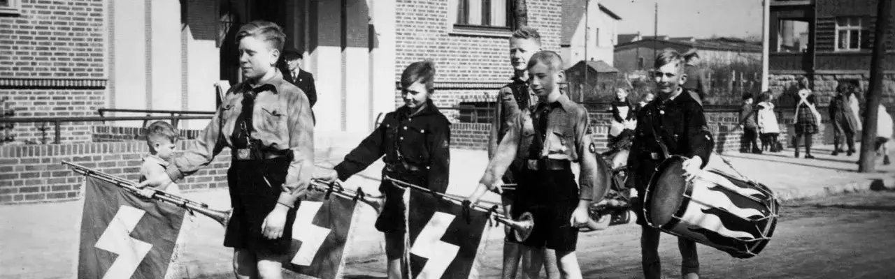 Jungen in Uniformen marschieren mit Fahnen und Trommeln. Sie repräsentieren die Ideologie der Volksgemeinschaft.
