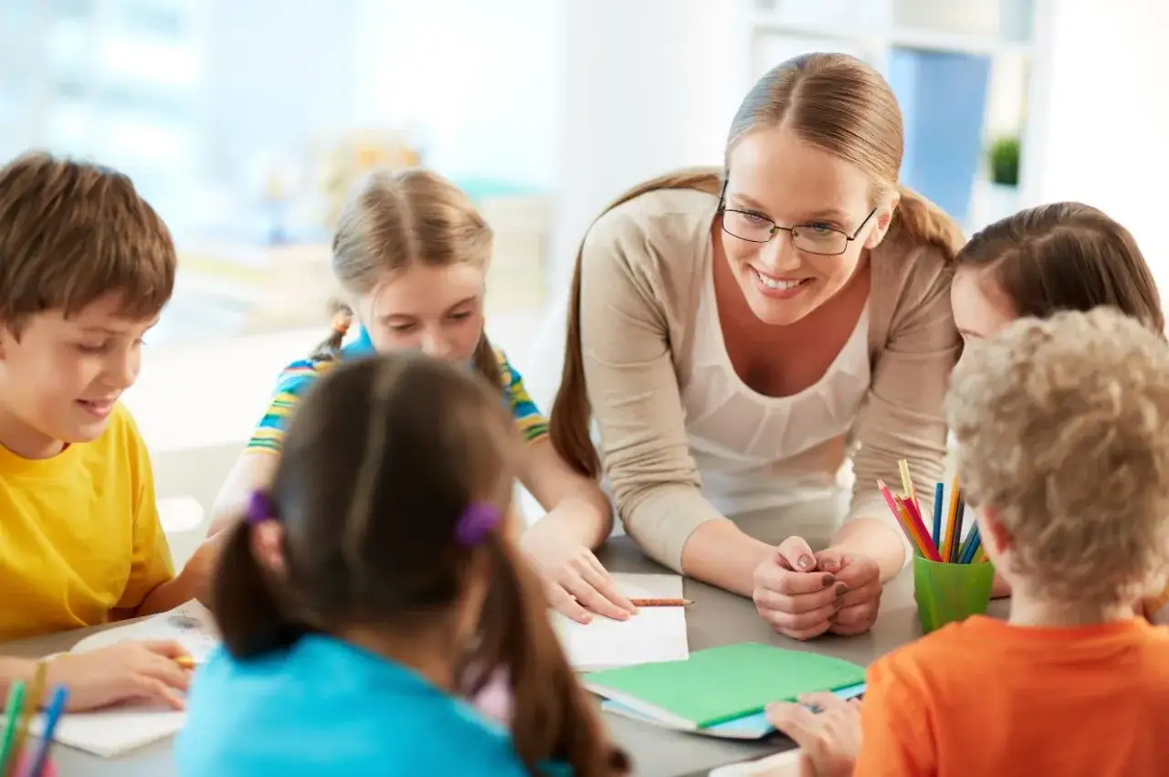 Lehrerin lächelt, während sie mit Kindern bei Kennenlernspielen am Tisch sitzt. Buntstifte und Hefte liegen bereit.