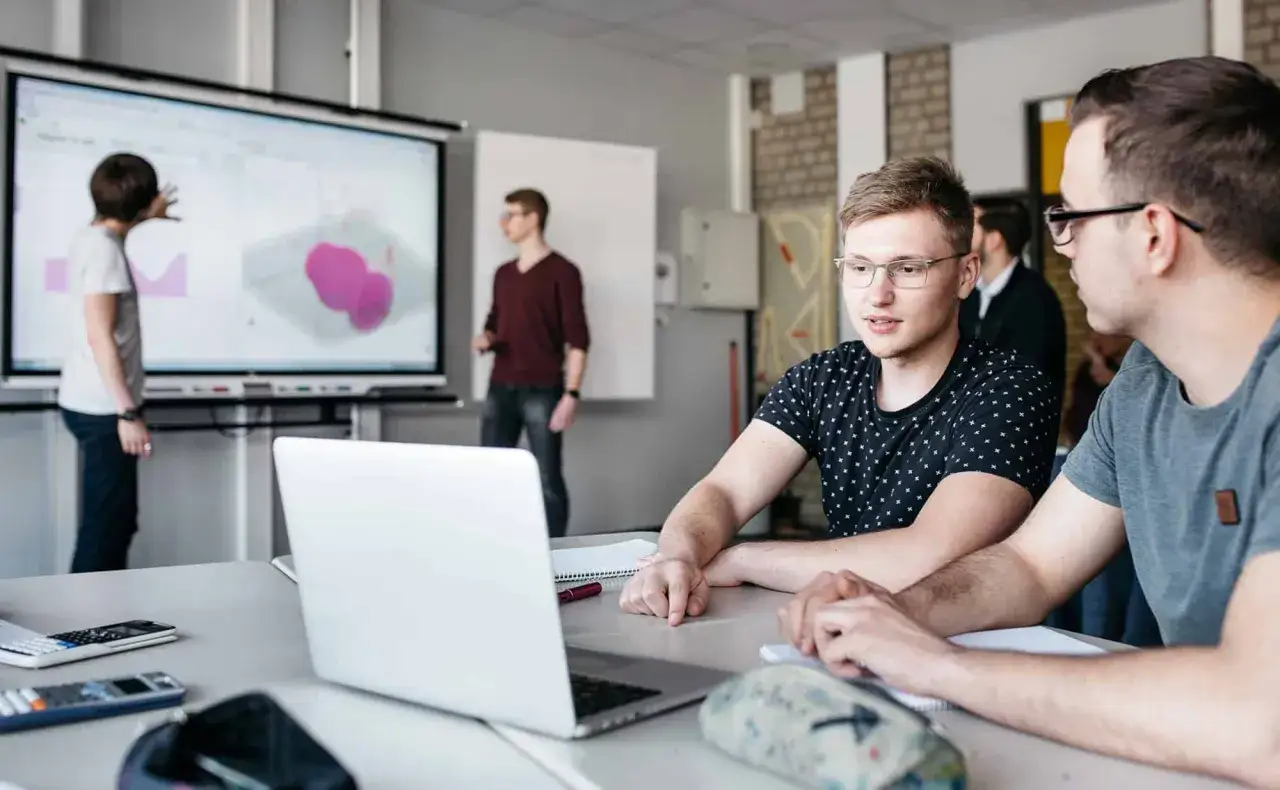 Zwei Studenten diskutieren über digitale Tools für den Unterricht, während ein Dozent an einem interaktiven Whiteboard erklärt.
