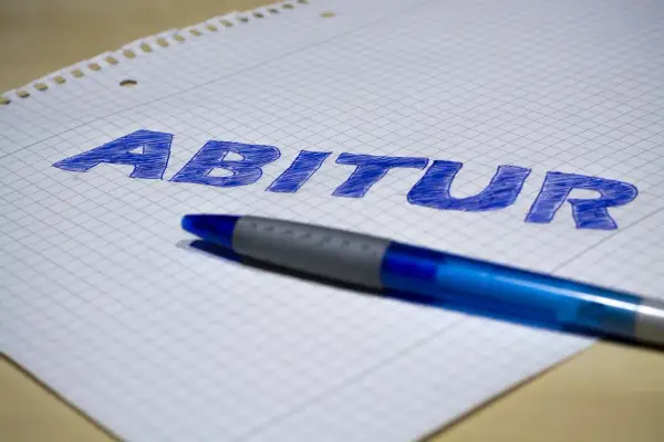 Blauer Stift liegt auf kariertem Papier, auf dem das Wort "Abitur" geschrieben ist. Der **Abitur**-Abschluss rückt näher.