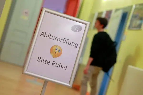 Schild mit der Bezeichnung "Abiturprüfung. Bitte Ruhe!" und einem Emoji, das zum Schweigen auffordert. Ein Schüler geht im Hintergrund vorbei.
