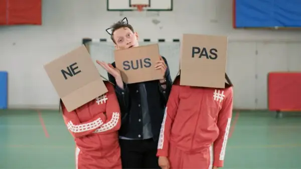 Drei Personen mit Pappkartons auf dem Kopf, auf denen "NE", "SUIS" und "PAS" steht. Sie bilden die französische Verneinung "ne suis pas".