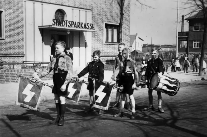 Jungen in Uniformen marschieren mit Fahnen und Trommel vor der Stadtsparkasse. Ein Bild der vermeintlichen Volksgemeinschaft.